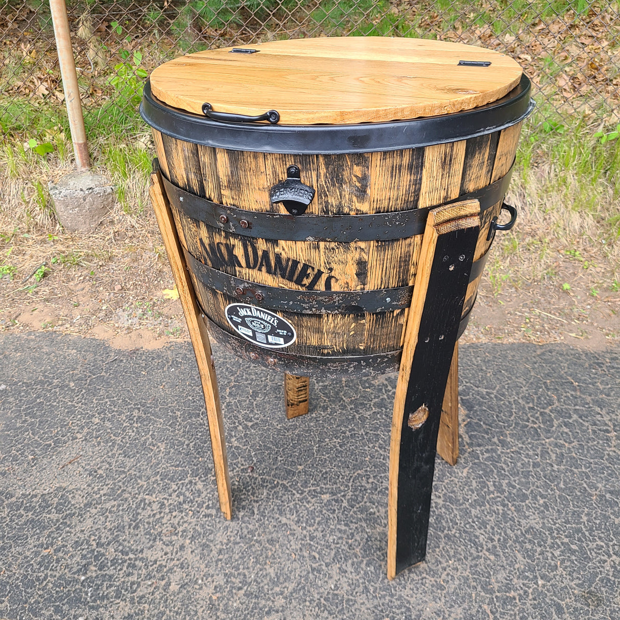 A Jack Daniel's Whiskey Barrel Cooler with Lid, priced at $395.00 (T008), exudes rustic charm as it stands on the pavement near a fence, repurposed as a standing BBQ grill.