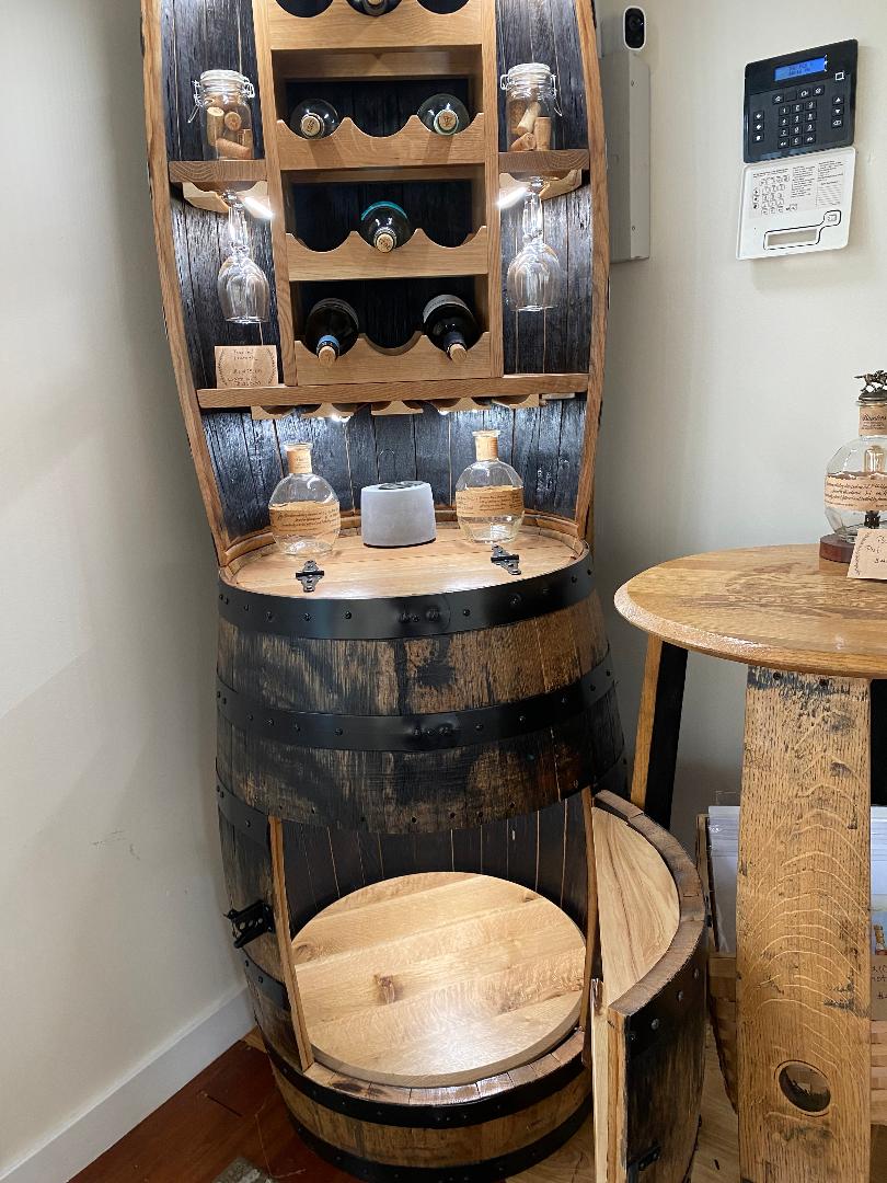 A Whiskey Barrel Hutch with Wine Rack and Lights, priced at $1750.00, featuring wine bottles and glasses, stands next to a small round wooden table adorned with decor items.