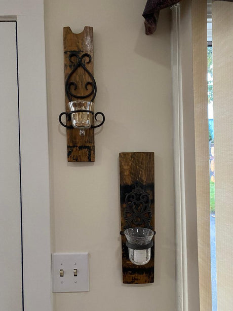 Two Whiskey Barrel Stave Candle Sconces with intricate designs and glass holders are mounted beside a light switch on a beige wall, crafted from reclaimed whiskey barrels. Priced at $75.00 each.