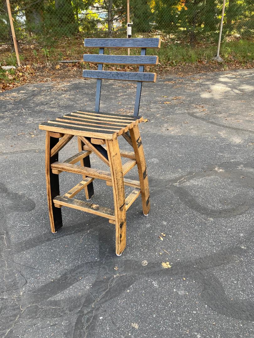 A Whiskey Barrel Stave Chair with Back, priced at $250.00, stands on an asphalt surface beside a chain-link fence and trees in the background.