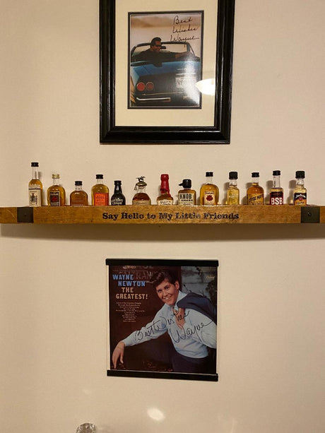 The Whiskey Barrel Stave Shelf For Nippers ($135.00), made from an old whiskey barrel, holds miniature liquor bottles. Above the shelf is a framed car photo, below it hangs a signed poster, and the setup is completed with the text "Say Hello to My Little Friends.