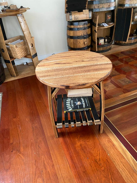 A Whiskey Barrel Stave Side Table with books on a shelf below, situated on a polished wood floor with whiskey barrels and shelves in the background. Price: $200.00.