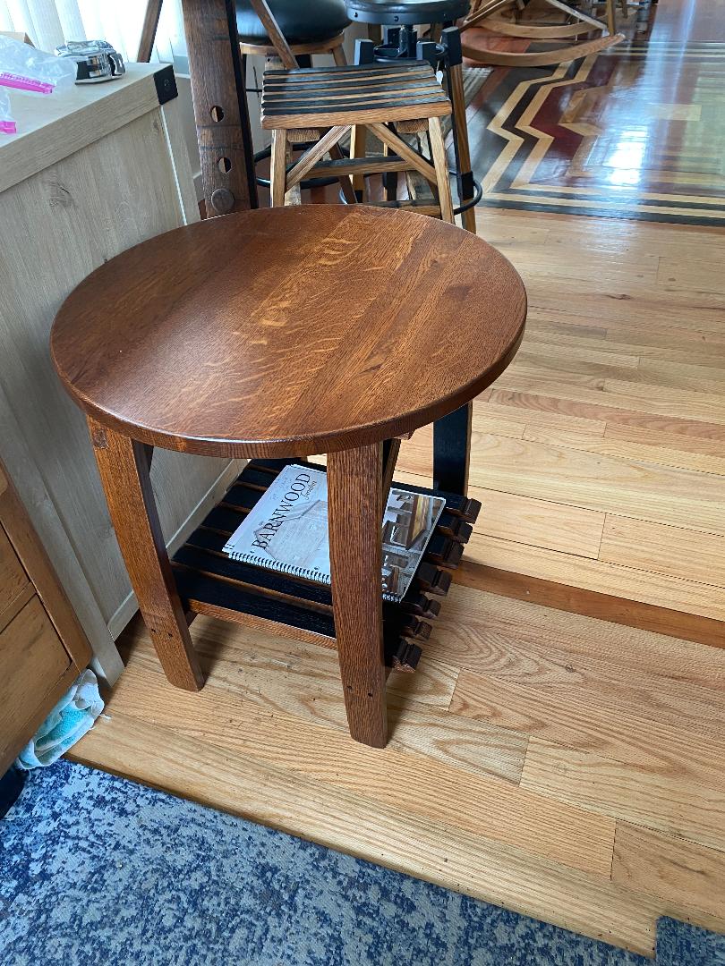 The Whiskey Barrel Stave Side Table, priced at $200.00, is a wooden round table with a lower shelf containing magazines, set on a hardwood floor next to a wooden chair.