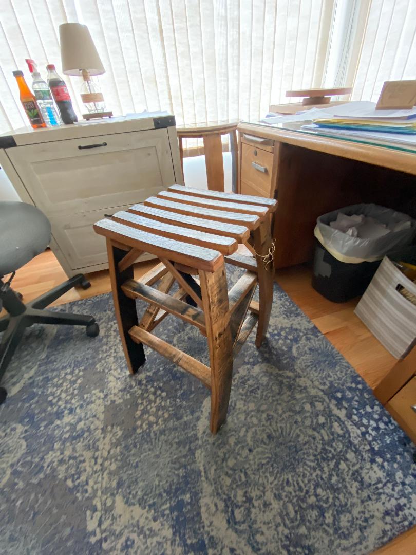 A Whiskey Barrel Stave Stool ($175.00) stands on a patterned blue rug between an office chair and a desk with various items in an office space.