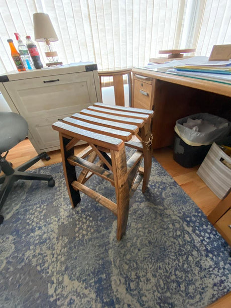 A Whiskey Barrel Stave Stool ($175.00) stands on a patterned blue rug between an office chair and a desk with various items in an office space.