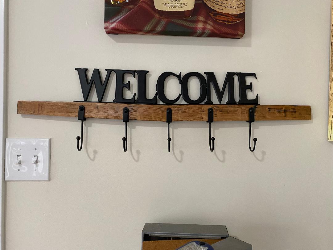 Whiskey Barrel Stave Welcome Coat Rack, crafted from reclaimed whiskey barrel wood with five hooks, mounted on a wall next to a light switch. $125.00