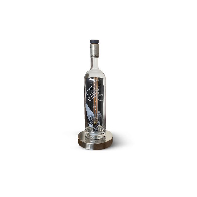 The Eagle Rare Lamp, featuring a tall glass bottle with clear liquid and a vintage design, is elegantly displayed on a round metallic stand.